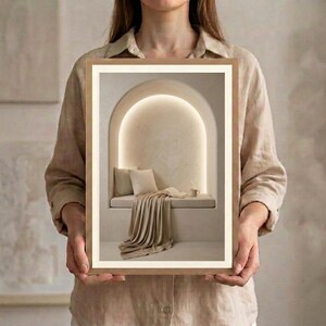 Hands holding a framed neutral reading nook wall art print in natural oak with a cream mat, showing the arched reading niche, soft cushions, and draped linen in warm beige tones.