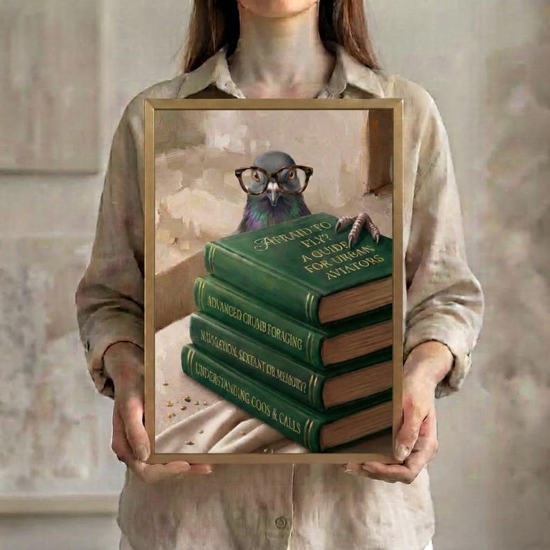 A person in a linen shirt holding the pigeon peeking over books art print in a slim gold frame against a soft neutral background