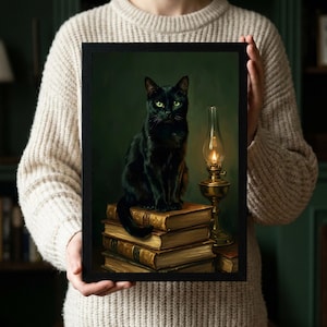 A person in a cream knit sweater holding a black framed print of a black cat sitting upright on old books beside a lit brass oil lamp.