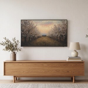 Spring orchard at dusk art from the set on a Frame TV in a neutral living room, above an oak credenza on legs with an olive tree, a lamp and books, in daylight from the left