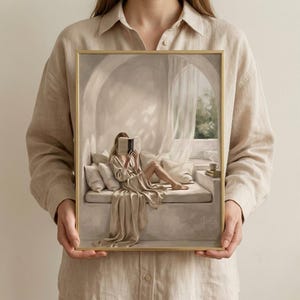 Person holding a framed neutral woman reading nook wall art print in a thin gold frame with no mat, showing the arched reading niche, draped linen, and warm beige tones.