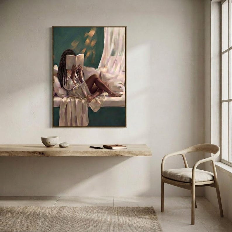 Gold-framed emerald green art print of a Black woman reading on a window seat above a floating oak shelf with two bowls and a woven chair in a bright minimal room.
