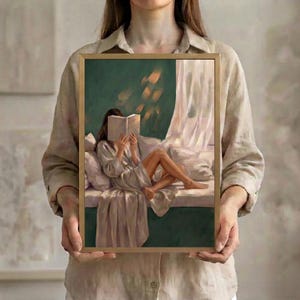 Woman holding a framed emerald green wall art print of a brunette woman reading in a window seat, showing true print size against a linen shirt.
