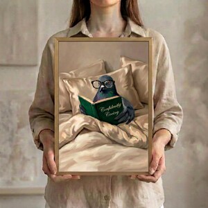 Person in a linen shirt holding the framed pigeon reading print in a slim gold frame, showing its true scale against a soft neutral background