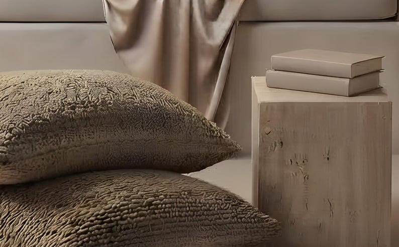 Close-up detail from Alcove Reading Wall Art, a cropped fragment of the printable artwork: a satin throw falling over the window seat, textured boucle floor cushions and a raw wood stool with two stacked books.