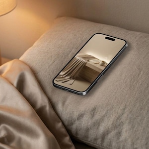 Puede incluir: Un tel&eacute;fono inteligente con marco oscuro y una pantalla que muestra una imagen en tonos beige de tela drapeada, un libro peque&ntilde;o y una taza. El dispositivo est&aacute; colocado en &aacute;ngulo sobre una almohada texturizada clara junto a una tela satinada.