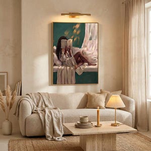 Gold-framed emerald green art print of a Black woman reading on a window seat above a cream sofa in a warm neutral living room, lit by a slim brass wall light, with candles on a low travertine table.