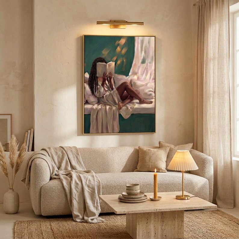 Gold-framed emerald green art print of a Black woman reading on a window seat above a cream sofa in a warm neutral living room, lit by a slim brass wall light, with candles on a low travertine table.