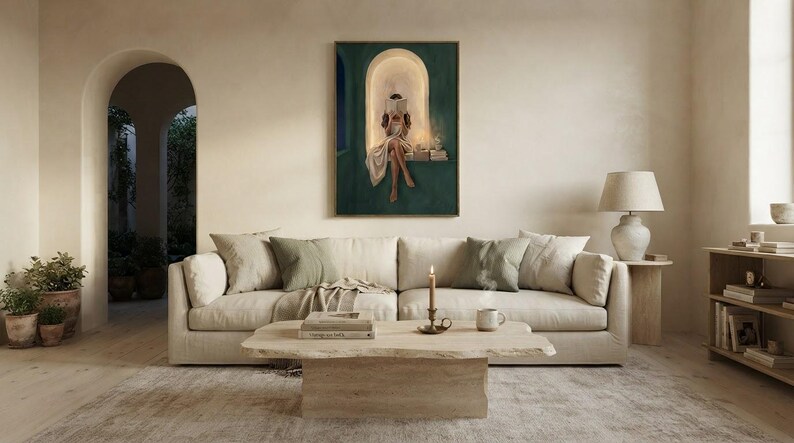 Emerald green reading nook art print above a deep cream sofa in an airy plaster living room, with a travertine coffee table, a lit candle and an arched doorway to a garden.