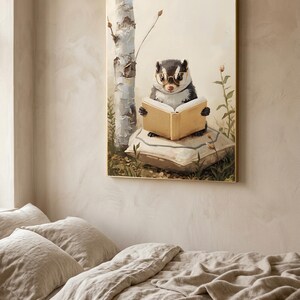 Framed woodland nursery print of a skunk in round glasses reading a book, hanging in a pale gold frame above a linen-covered bed in a warm neutral bedroom