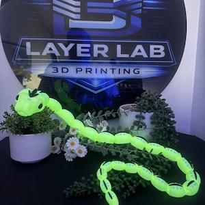 Puede incluir: Un juguete de serpiente articulada de color amarillo neón con marcas negras y una pequeña cresta en la cabeza. La serpiente tiene un cuerpo segmentado que se curva sobre la superficie. El fondo muestra un letrero con LAYER LAB 3D PRINTING.