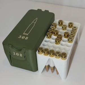 May include: An olive green rectangular storage box with a white line drawing of a bullet and the text 308 printed on the lid and front. Beside it is a white plastic ammunition tray holding multiple brass-cased cartridges in an upright position.