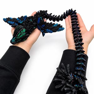 May include: A black articulated dragon toy with blue and green accents. The dragon is held in two hands, with the tail extending down the arm. The dragon's wings and body have a textured, layered design.