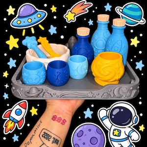 May include: A grey tray with space-themed relief designs holds a collection of silicone items including two blue bottles with caps, a white bowl with a star-shaped tool, and four small cups in blue and yellow featuring alien, sun, and moon motifs.