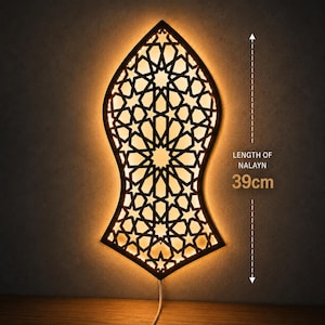 Puede incluir: Una lámpara de pared con forma de Nalayn, con marco negro y un intrincado patrón geométrico de estrellas. La lámpara emite un brillo cálido y suave a través del diseño recortado. El texto indica una longitud de 39 cm para el Nalayn.