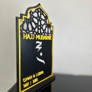 May include: A black and gold decorative plaque with an arched top featuring an intricate geometric lattice pattern. The text reads Hajj Mubarak, Omar & Lubna, and 1447 / 2026. A white Arabic calligraphic symbol is centred on the black surface.