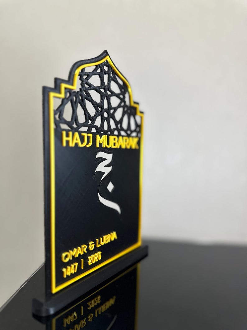 May include: A black and gold decorative plaque with an arched top featuring an intricate geometric lattice pattern. The text reads Hajj Mubarak, Omar & Lubna, and 1447 / 2026. A white Arabic calligraphic symbol is centred on the black surface.