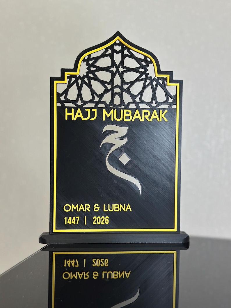 May include: Black and gold Hajj Mubarak plaque with geometric detailing. The plaque reads "HAJJ MUBARAK" and features the names "OMAR & LUBNA" with the dates 1447 | 2026. The plaque is displayed on a black stand.