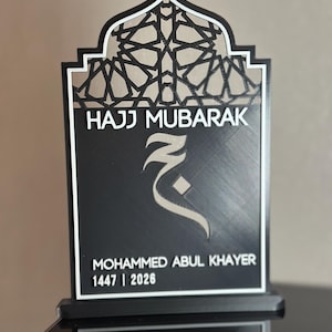 May include: A black and white plaque featuring an arched top with a geometric lattice pattern. The text HAJJ MUBARAK is printed above a stylised Arabic letter. Below, the name MOHAMMED ABUL KHAYER and the numbers 1447 | 2026 are displayed.