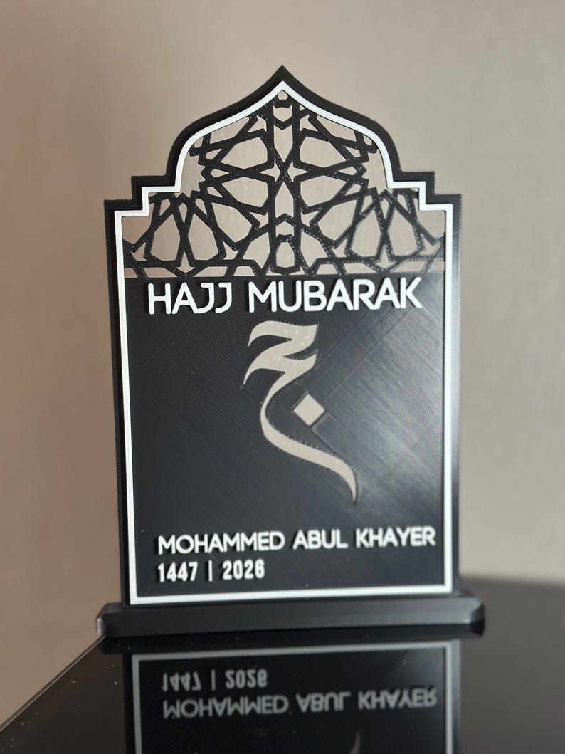 May include: A black and white plaque featuring an arched top with a geometric lattice pattern. The text HAJJ MUBARAK is printed above a stylised Arabic letter. Below, the name MOHAMMED ABUL KHAYER and the numbers 1447 | 2026 are displayed.