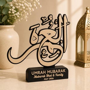 May include: A decorative tabletop sign featuring gold Arabic calligraphy on a black base. The base displays the text "UMRAH MUBARAK", "Mubarak Bhai & Family", and "JULY 2026" in gold lettering. The structure is composed of a black frame with gold accents.