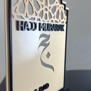 May include: A beige and black 3D-printed plaque with an arched top featuring an intricate geometric pattern. The text reads Hajj Mubarak, Mom & Dad, and 1447 / 2026. A stylised Arabic letter is centred on the plaque, which stands on a black base.