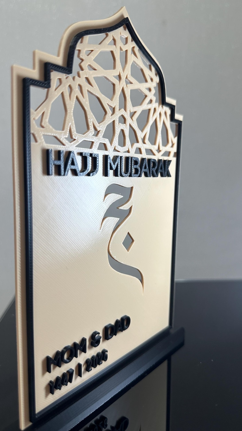 May include: A beige and black 3D-printed plaque with an arched top featuring an intricate geometric pattern. The text reads Hajj Mubarak, Mom & Dad, and 1447 / 2026. A stylised Arabic letter is centred on the plaque, which stands on a black base.