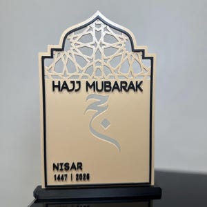 May include: A beige and black decorative plaque featuring an arched top with a geometric lattice pattern. The text reads HAJJ MUBARAK, followed by an Arabic character, the name NISAR, and the years 1447 | 2026. The plaque stands on a black base.