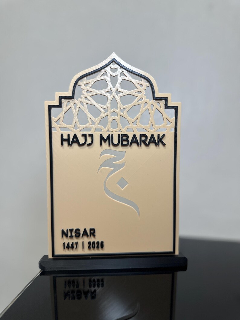May include: A beige and black decorative plaque featuring an arched top with a geometric lattice pattern. The text reads HAJJ MUBARAK, followed by an Arabic character, the name NISAR, and the years 1447 | 2026. The plaque stands on a black base.