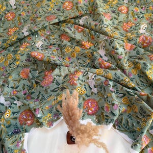 May include: A sage green fabric featuring a repeating floral pattern with pink, orange, and yellow blossoms, along with green leaves and white accents. The textile is draped in soft folds, showcasing the intricate botanical print across the surface.