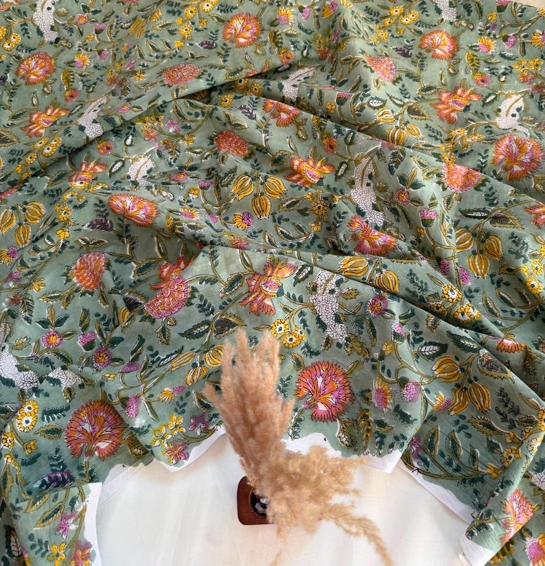 May include: A sage green fabric featuring a repeating floral pattern with pink, orange, and yellow blossoms, along with green leaves and white accents. The textile is draped in soft folds, showcasing the intricate botanical print across the surface.