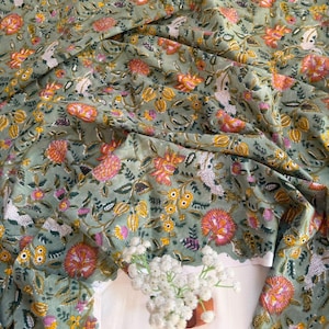 May include: A sage green fabric featuring a repeating floral pattern with orange, pink, and yellow blossoms, along with small white textured clusters and dark green leaves. The textile is draped in soft folds, displaying a dense botanical print.