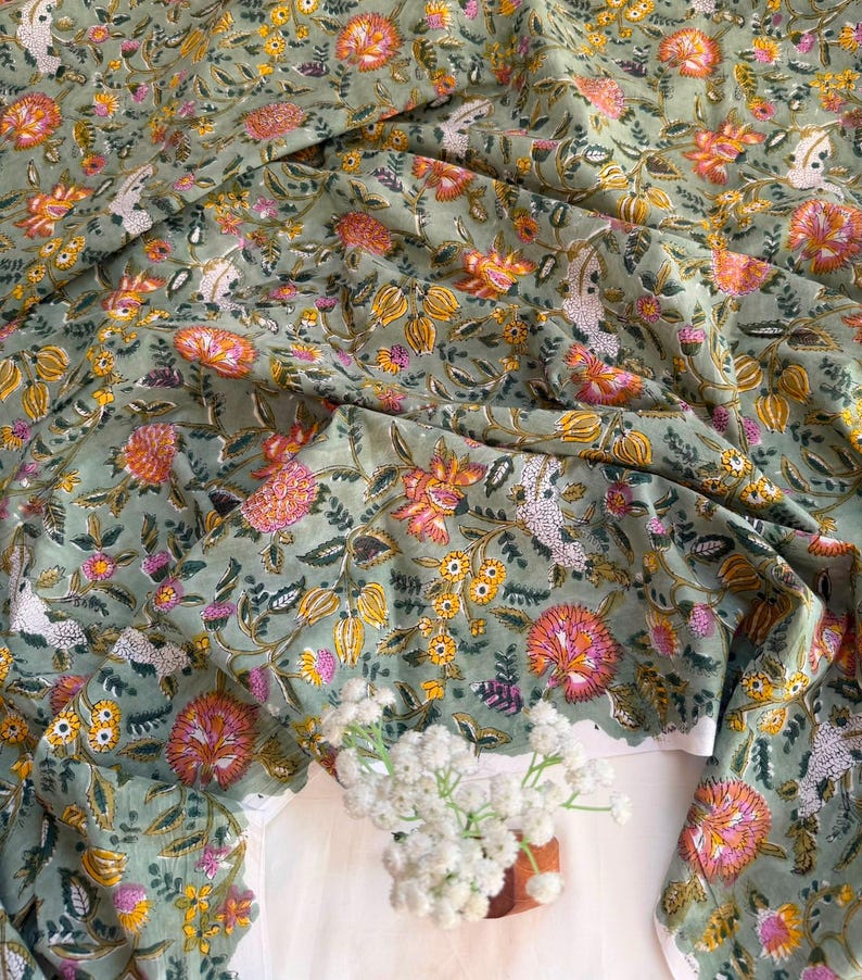 May include: A sage green fabric featuring a repeating floral pattern with orange, pink, and yellow blossoms, along with small white textured clusters and dark green leaves. The textile is draped in soft folds, displaying a dense botanical print.