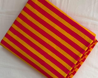 May include: A folded piece of fabric featuring a repeating pattern of bold, diagonal stripes in alternating shades of vibrant magenta pink and bright golden yellow. The textile is neatly stacked, showcasing the consistent width of the stripes.