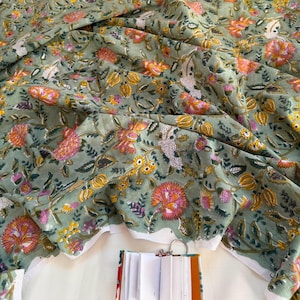 May include: A sage green fabric featuring a repeating floral pattern in shades of orange, pink, and yellow with green leaves. A small, open, white-paged notebook with a colorful spine and a metal ring binder is positioned at the bottom center.