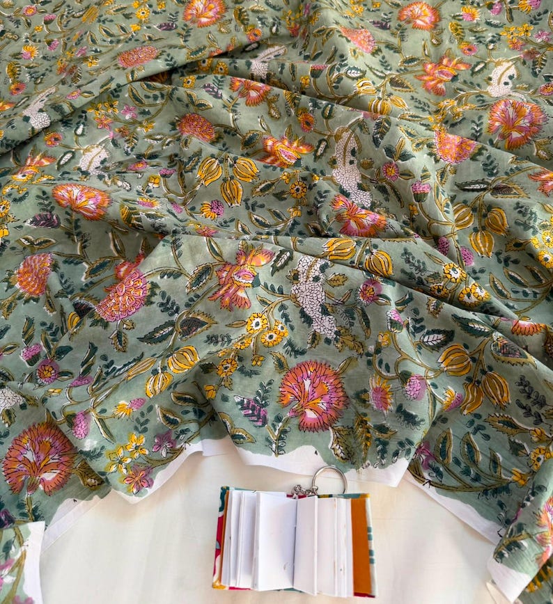 May include: A sage green fabric featuring a repeating floral pattern in shades of orange, pink, and yellow with green leaves. A small, open, white-paged notebook with a colorful spine and a metal ring binder is positioned at the bottom center.