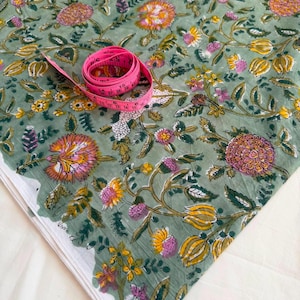 May include: A sage green fabric featuring a repeating floral pattern in shades of pink, yellow, and white, accompanied by a coiled pink measuring tape showing markings in inches and centimeters to indicate the scale of the textile design.