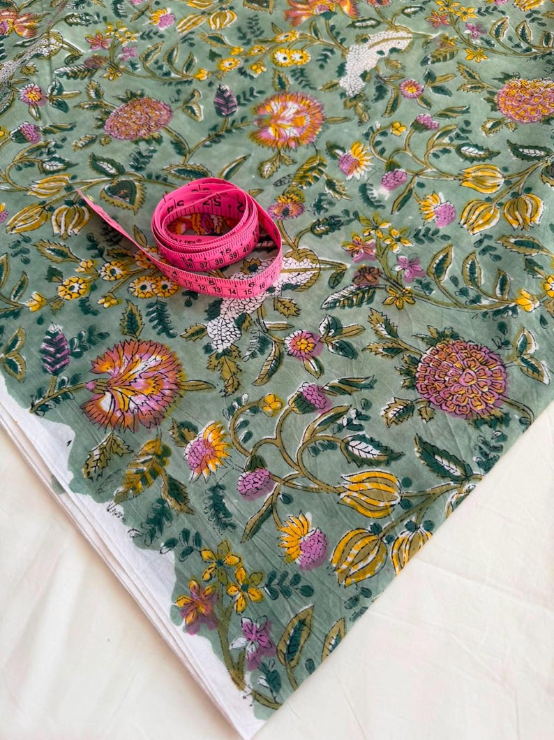May include: A sage green fabric featuring a repeating floral pattern in shades of pink, yellow, and white, accompanied by a coiled pink measuring tape showing markings in inches and centimeters to indicate the scale of the textile design.