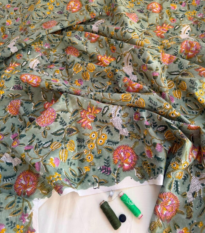 May include: A sage green fabric featuring a repeating floral pattern in shades of pink, orange, and yellow. The textile is draped to show folds and texture. Included are two spools of thread in dark green and bright green, and one small black button.
