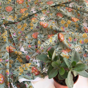 May include: A sage green fabric featuring a repeating floral pattern with orange, yellow, and pink blossoms, along with green leaves and white bird motifs. The textile is draped in soft folds, showcasing the intricate botanical print across the surface.
