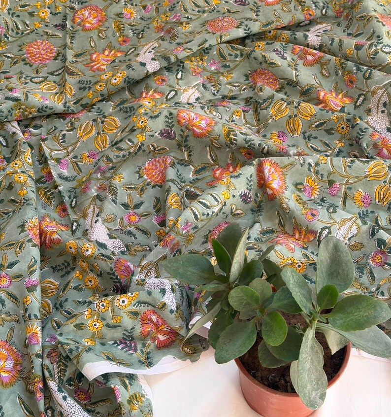 May include: A sage green fabric featuring a repeating floral pattern with orange, yellow, and pink blossoms, along with green leaves and white bird motifs. The textile is draped in soft folds, showcasing the intricate botanical print across the surface.
