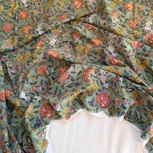 May include: A sage green fabric featuring a dense floral pattern with orange, yellow, and white blossoms, along with green leaves and stems. The textile displays a repeating botanical print with intricate details, arranged in a flowing, gathered drape.