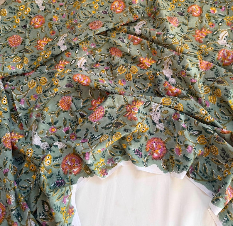 May include: A sage green fabric featuring a dense floral pattern with orange, yellow, and white blossoms, along with green leaves and stems. The textile displays a repeating botanical print with intricate details, arranged in a flowing, gathered drape.