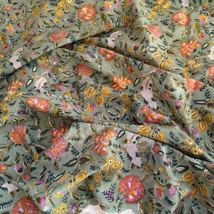 May include: A sage green fabric featuring a repeating floral pattern with orange, yellow, and pink blossoms, along with dark green leaves and stems. The textile is draped in soft folds, highlighting the intricate botanical print across the surface.