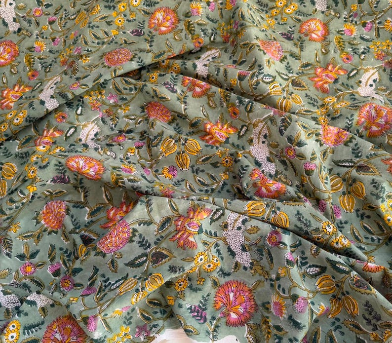 May include: A sage green fabric featuring a repeating floral pattern with orange, yellow, and pink blossoms, along with dark green leaves and stems. The textile is draped in soft folds, highlighting the intricate botanical print across the surface.