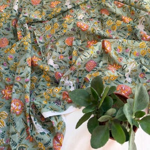May include: A sage green fabric featuring a repeating floral pattern with orange, yellow, and white blossoms and green leaves. The textile has a soft, draped appearance with folds, and a small potted green plant sits in the lower right corner.