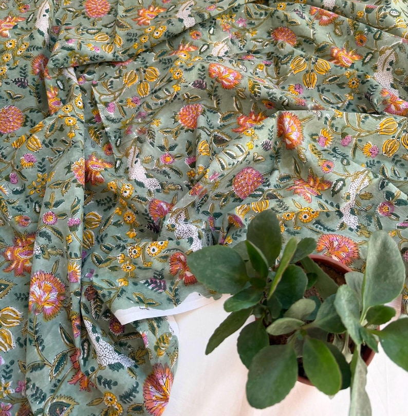 May include: A sage green fabric featuring a repeating floral pattern with orange, yellow, and white blossoms and green leaves. The textile has a soft, draped appearance with folds, and a small potted green plant sits in the lower right corner.