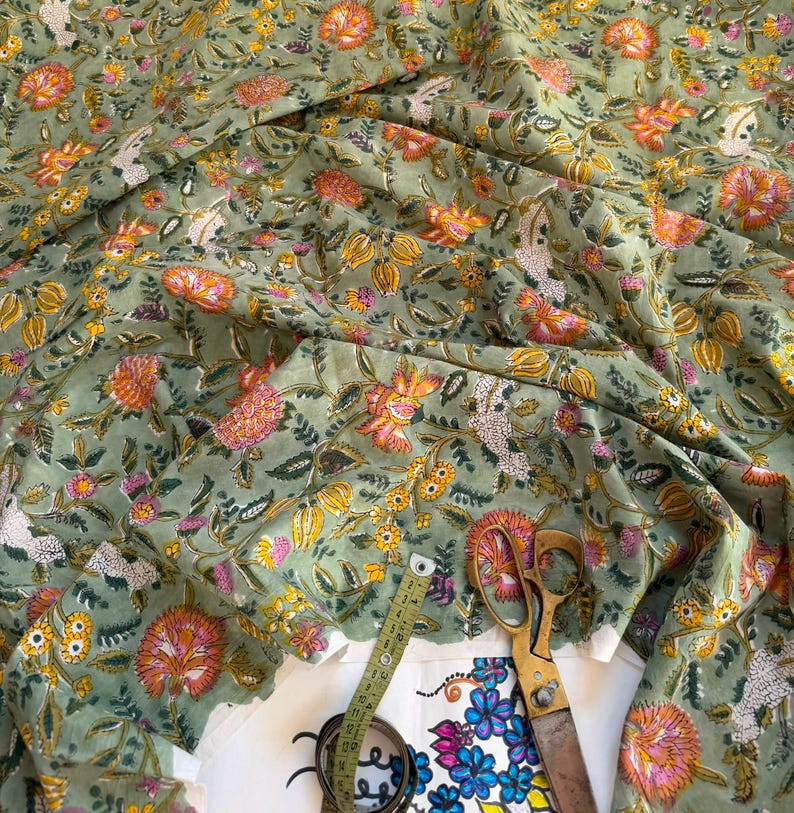 May include: A sage green fabric featuring a repeating floral pattern in shades of orange, yellow, and pink. A yellow measuring tape is positioned over the fabric, showing a measurement of approximately 12 inches along the visible length of the tape.
