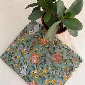 May include: A folded piece of fabric featuring a sage green background with a repeating floral pattern in shades of orange, yellow, pink, and white. The textile displays intricate botanical illustrations of various flowers and leaves.