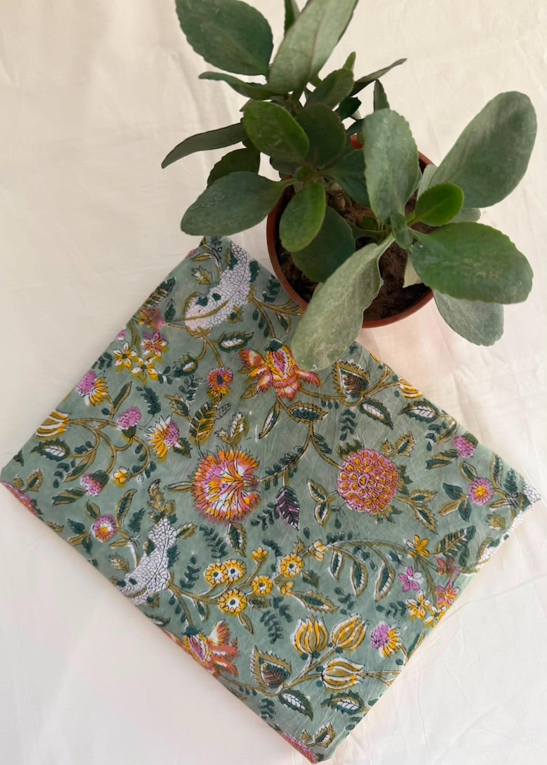May include: A folded piece of fabric featuring a sage green background with a repeating floral pattern in shades of orange, yellow, pink, and white. The textile displays intricate botanical illustrations of various flowers and leaves.